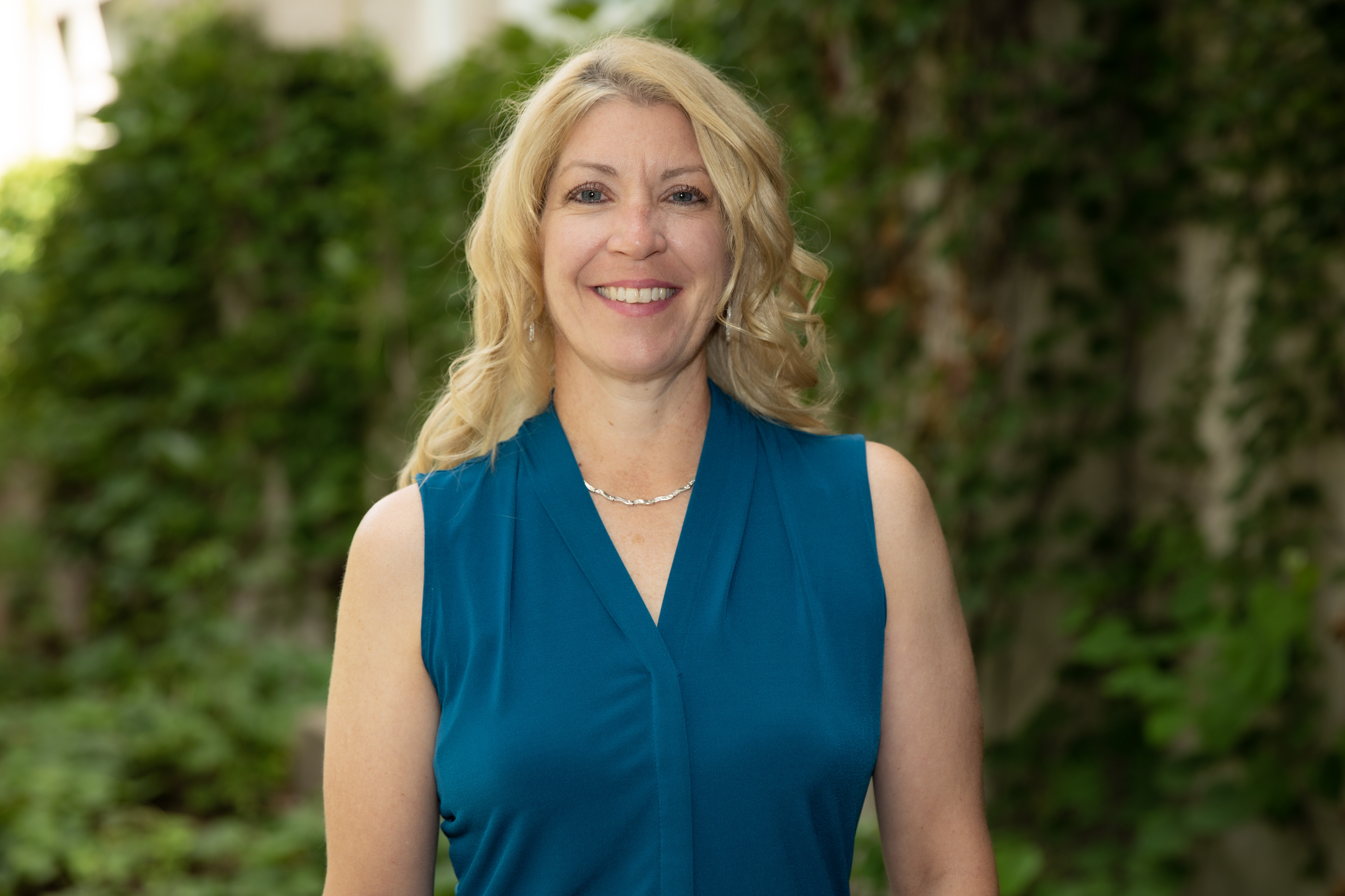 A person with long, wavy blonde hair wearing a sleeveless teal blouse, smiling while standing outdoors with greenery in the background.