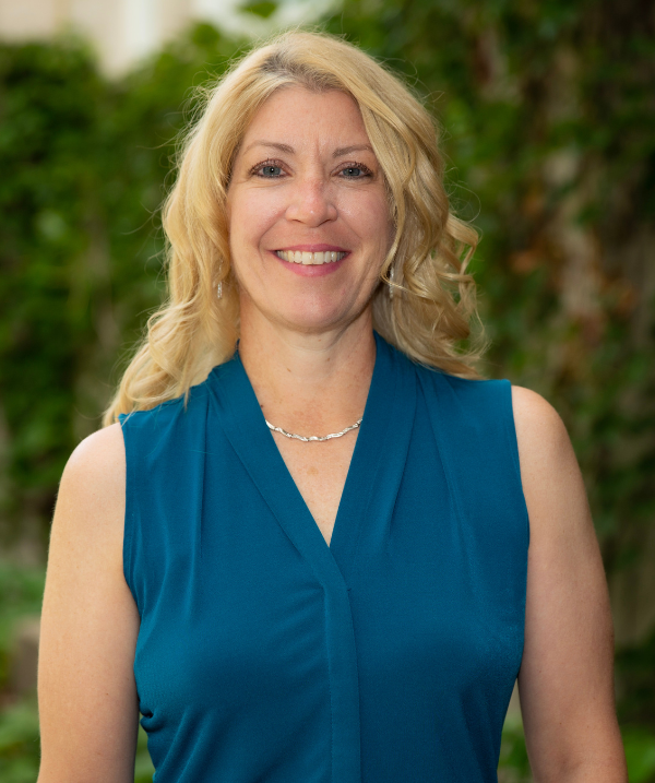 Tracey Covassin headshot. Tracey smiles for the camera while standing outside of IM Sport Circle, home of the Department of Kinesiology. Covassin wears a dark blue, sleeveless top with a silver necklace. Covassin's blonde hair is curled and falls past the shoulders.