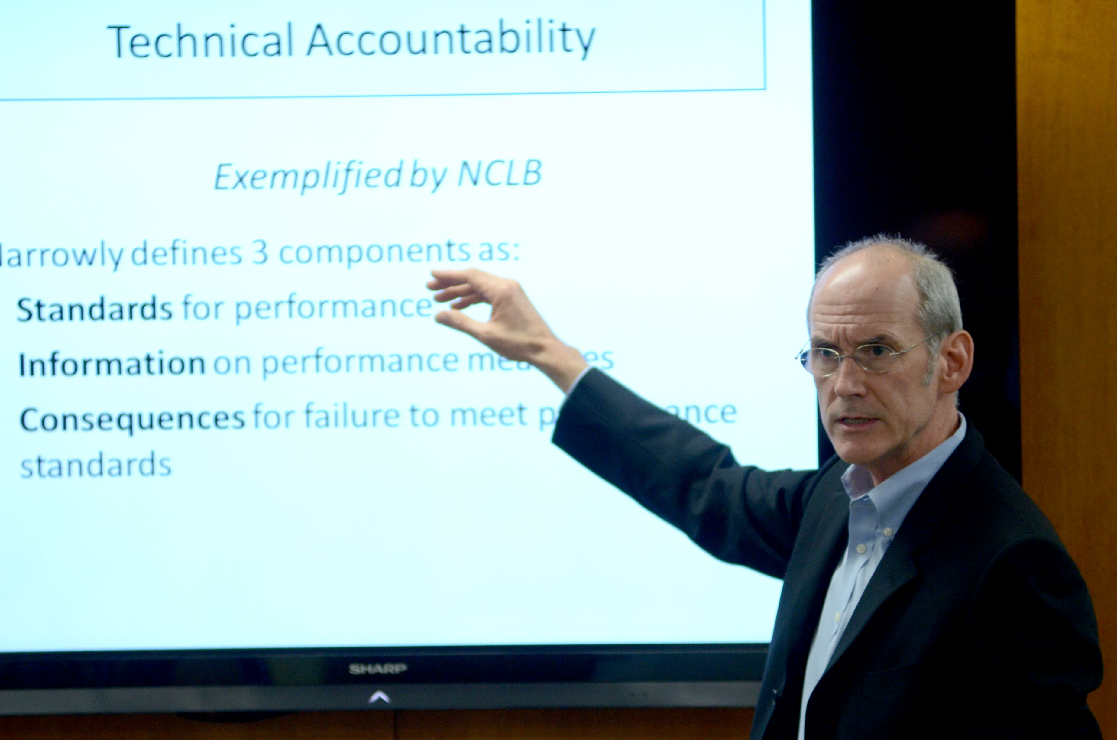Arsen speaks during a presentation, lifting the right arm to point at a PowerPoint slide on a screen. Arsen wears a white button-down shirt under a black blazer and wire-rimmed glasses. Arsen's gray hair is short.
