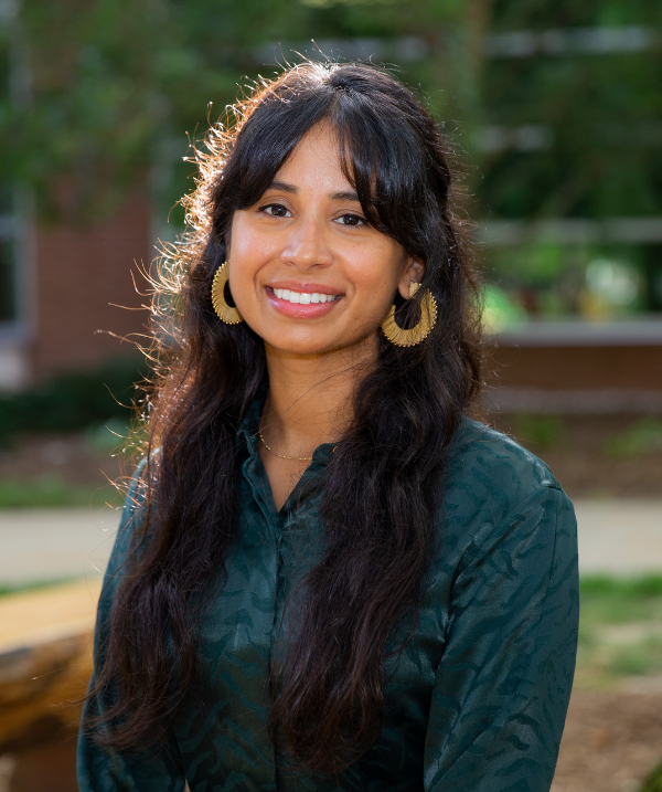 Tamsinda Dhaliwal smiles toward the camera in an outdoor setting. Dhaliwal wears a dark-green button-down blouse and large gold hoop earrings. Dhaliwal's hair is black and falls to mid-torso.