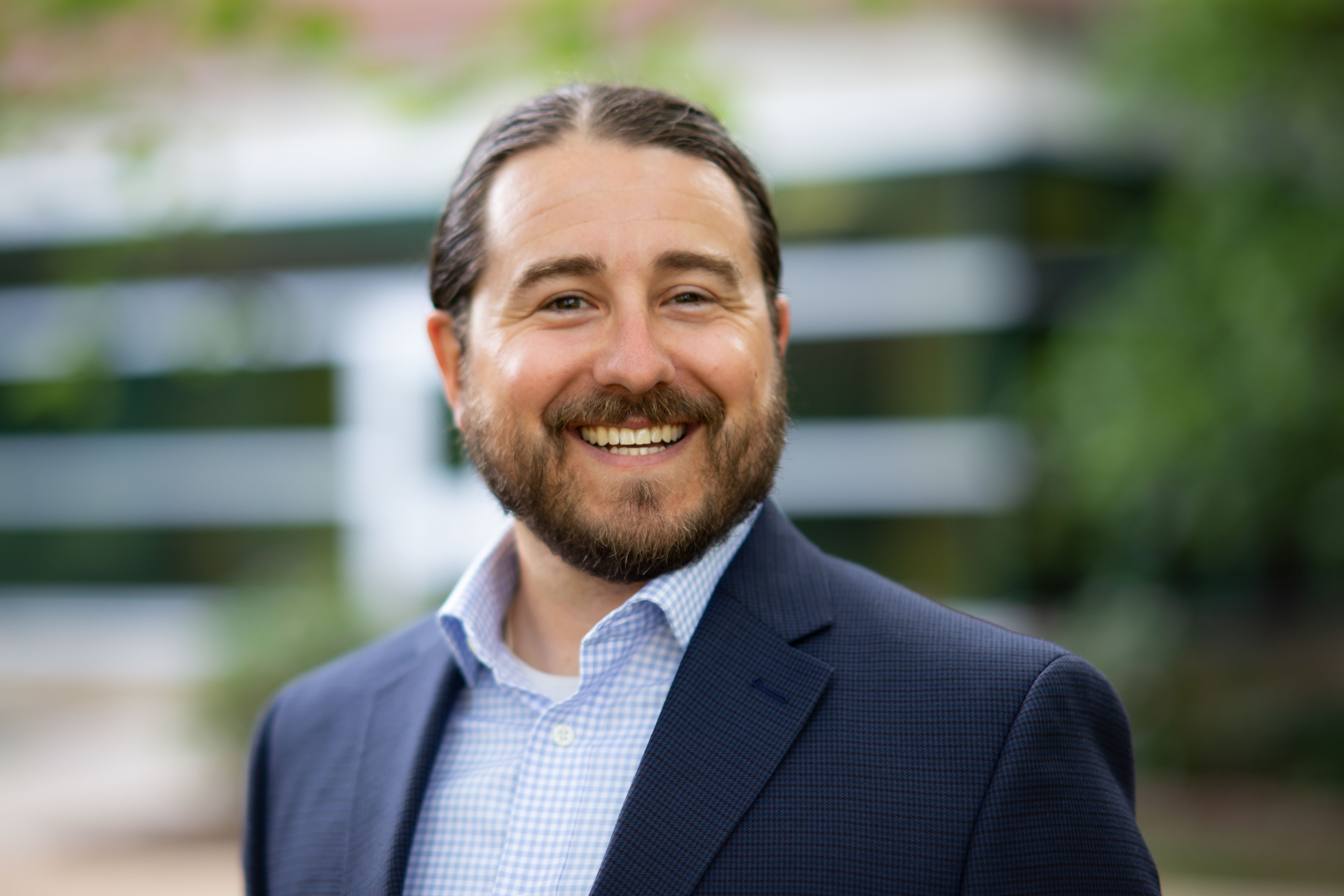 Ryan Smith wearing a navy blue suit jacket and light blue shirt. There is a glass and brick building out of focus in the background. 