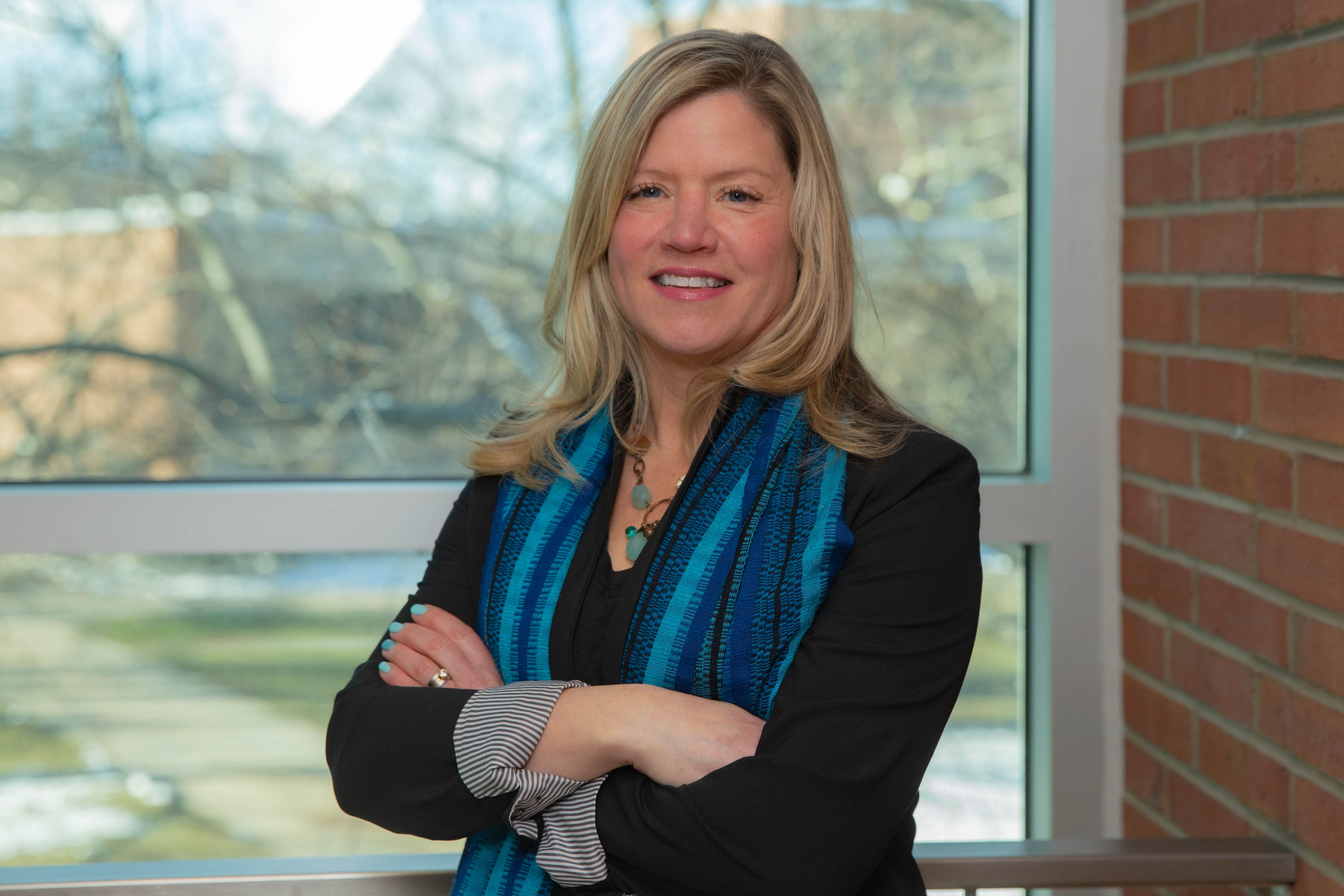 Beth Herbel-Eisenmann wearing a dark jacket with a blue scarf. She is standing in front of a window. 
