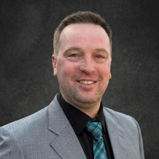 Brad Cox wearing a gray suit jacket, black shirt and blue patterned tie. 