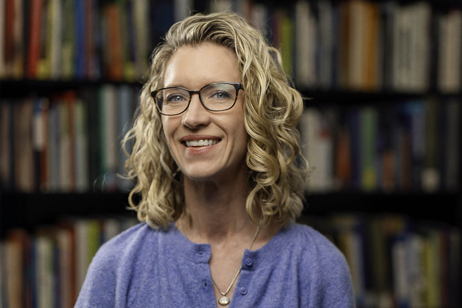Kristen Bieda wearing a blue blouse and glasses. There is a bookshelf full of books in the background.  