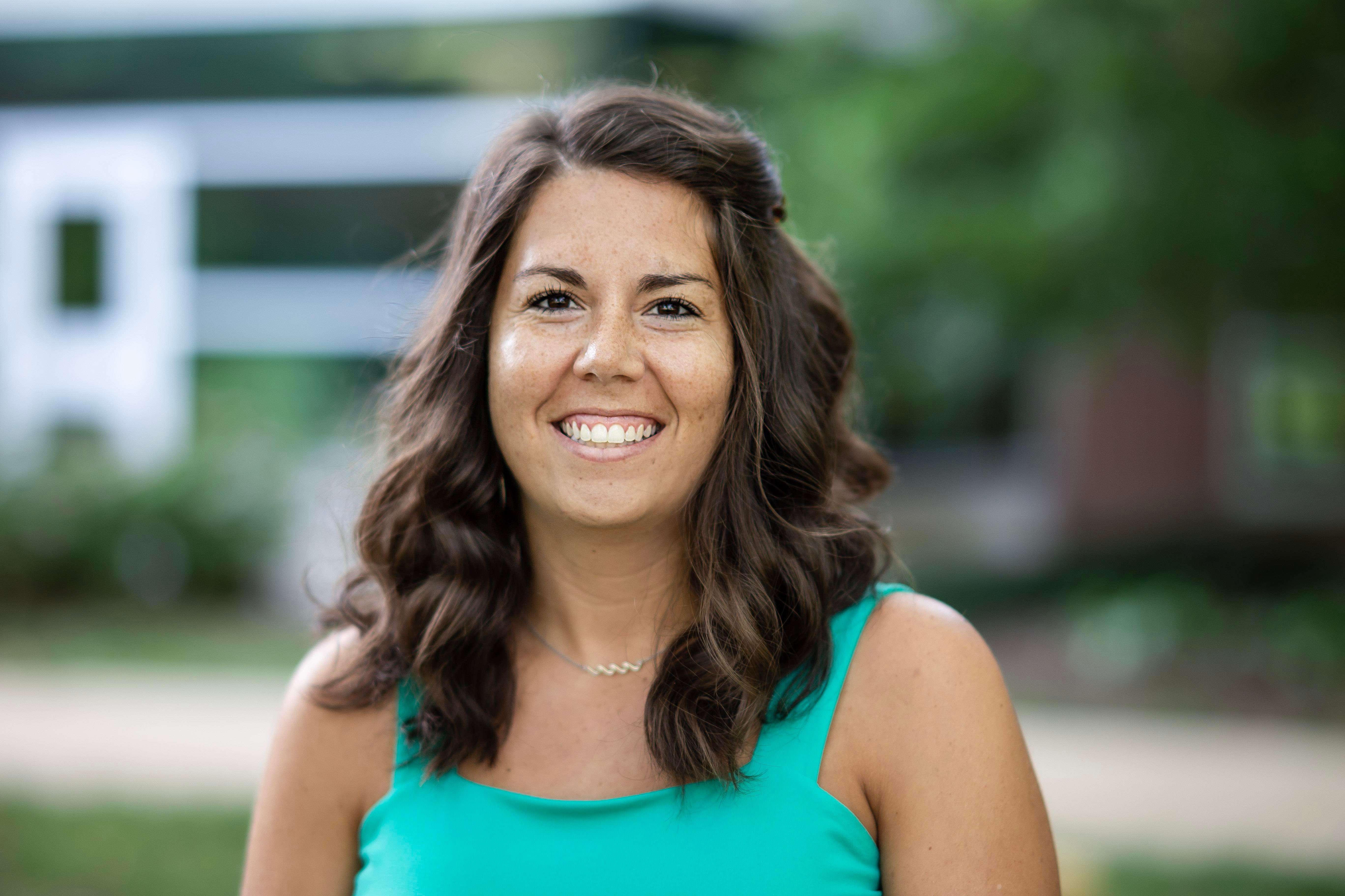Ashley Bingel wearing a teal top. There is a glass and brick building out of focus in the background. 