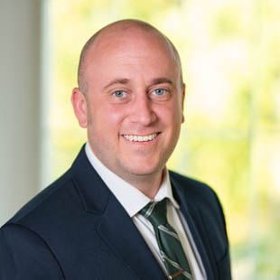 Headshot of William Adams wearing a dark suit and green tie