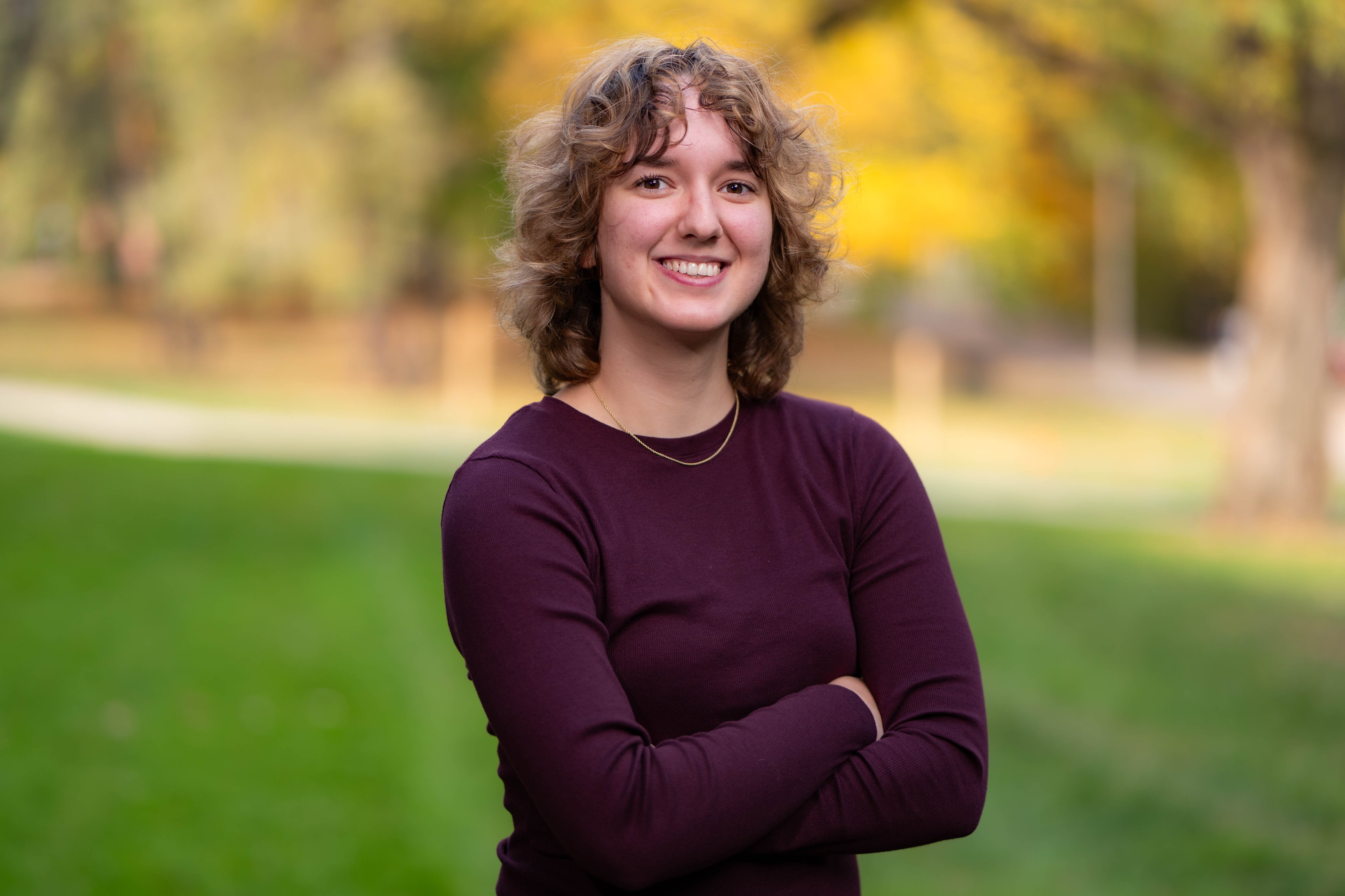 Adria Mestré wearing a burgandy sweater. There is green foliage in the background. 