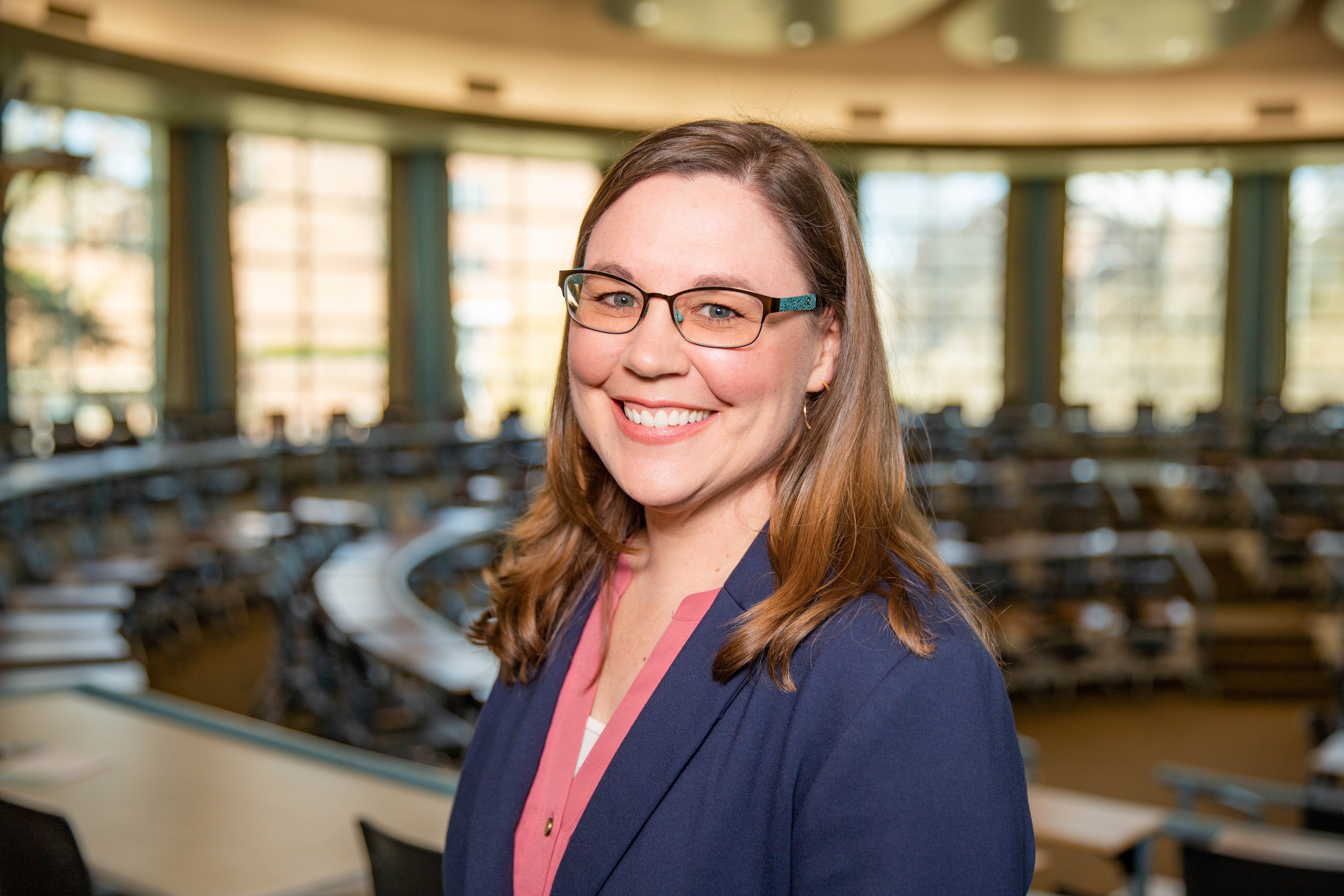 Alicia Strain wearing a navy jacket, with a pink blouse and glasses. She is standing in the KIVA. 
