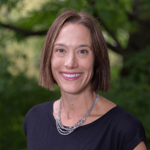 Jana Aupperlee wearing a dark blue shirt with a silver necklace. There is greenery in the background.