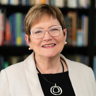 Ann Austin headshot. Austin poses against a bookshelf. Austin wears a black blouse under a beige blazer. Austin wears a necklace with a large, silver, circular pendant. Austin's blondeish hair is short. Austin wears glasses and earrings.