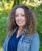 Christina Berchini Wyman smiles for the camera in an outdoor setting. Wyman wears a navy blue top under a denim jacket. Wyman's hair is brown and curly.