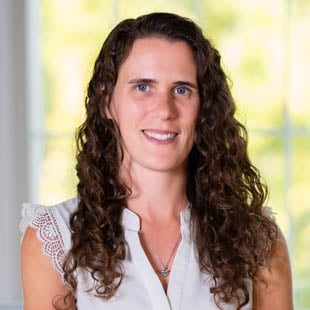 Headshot of Amy Boettcher wearing a white blouse. She is standing in front of windows with greenery in the background.