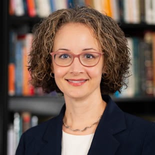 Headshot of Kristine Bowman wearing a dark jacket, white shirt and glasses with a bookshelf in the background.