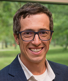 Matthew Brodhead. He wears a navy blazer over a white button down shirt. His dark hair is cropped short. He wears dark-rimmed glasses.