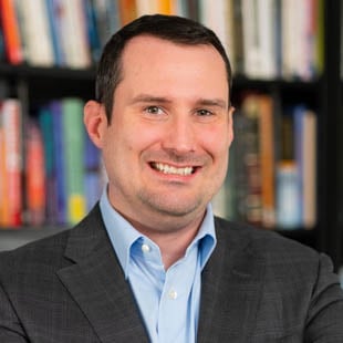 Headshot of Jason Burns wearing a dark suit jacket and light blue shirt, standing in front of a bookshelf
