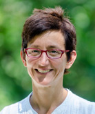Angela Calabrese Barton smiles for the camera in an outdoor setting, standing in front of a bush lush with green leaves. Barton wears a white shirt and red-rimmed glasses. Barton has short, dark brown hair.