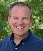 John Carlson smiles in an outdoor setting in front of a bush lush with green leaves. Carlson wears a dark blue button-down shirt. Carlson's blonde and gray hair is short. 