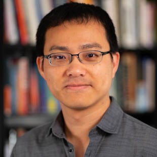 Headshot of Wei-Chu Chen wearing a dark shirt and glasses, standing in front of a bookshelf