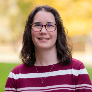 Kristy Chene wearing a red and white stripe sweater and glasses. There is fall foliage in the background. 