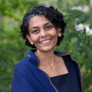 Headshot of Amita Chudgar wearing a blue jacket and blue shirt. She is standing in front of green foliage.
