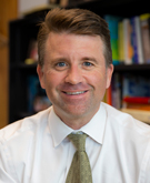 Portrait of a smiling man with short, light brown hair wearing a white dress shirt and a green patterned tie, sitting in an office with bookshelves filled with books in the background."