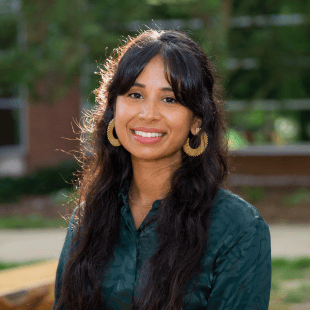 Tasmin Dhaliwal wearing a dark green shirt and gold earrings. There is a brick and glass building in the background. 