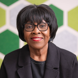 Patricia Edwards wearing a dark jacket, dark shirt and glasses. She is standing in front of a wall with a green hexagon pattern. 