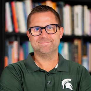 Scott Farver wearing a green polo with a white Spartan helmet and glasses in front of a bookcase