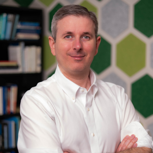 Headshot of David Ferguson wearing a white button shirt. He is standing in front of bookshelves and a wall with a green hexagon pattern. 