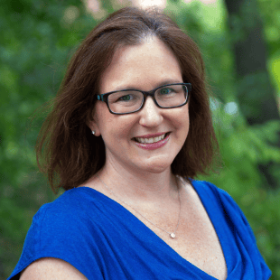 Amelia Gotwals Wenk wearing a blue shirt and glasses. There are green trees in the background. 