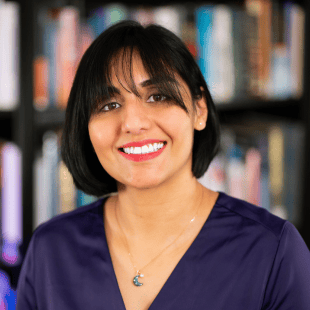 Harvi Hart wearing a purple blouse and a blue necklace. There are bookshelves in the background. 