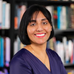 Headshot of Harvi Hart standing in front of a bookshelf. She is wearing a purple blouse.
