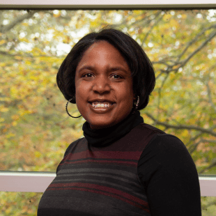 Latisha Hurd wearing a brown and red sweater. There is fall foliage in the background. 