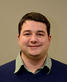 Scott Imberman smiles for the camera against a neutral beige background. Scott wears a green button-down under a dark blue sweater. Scott has dark brown, short hair.