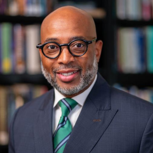 Jerlando F. L. Jackson headshot. Jackson poses in front of a bookshelf. Jackson wears a dark blazer over a green tie. Jackson has dark-rimmed glasses on.
