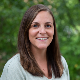 Headshot of Tiffany Joy wearing a light blue shirt, standing in front of green foliage