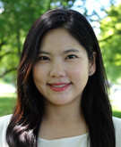 A portrait of a woman with long dark hair, smiling outdoors with green trees and sunlight in the background. She is wearing a light-colored blouse and has a friendly expression.