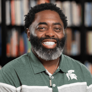 Curtis Lewis wearing a green and gray Spartan polo. There is a bookshelf in the background. 