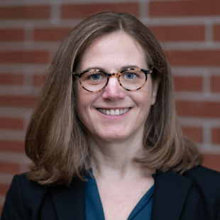 Lisa Linnenbrink-Garcia wearing a dark jacket, blue shirt and glasses. She is standing in front of a brick wall.
