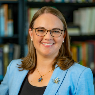 Headshot of Madeline Mavrogordato wearing a light blue jacket, black shirt and glasses. There is a bookshelf in the background