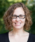 Amanda McNew. She wears a black, short-sleeved blouse. Her auburn hair is curly and cropped above her shoulders. She wears dark-rimmed glasses. 