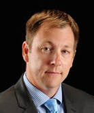 Andrew Nay headshot. Andrew wears a light blue button down shirt and a complementary tie with a black blazer. Andrew's hair is cropped short. 