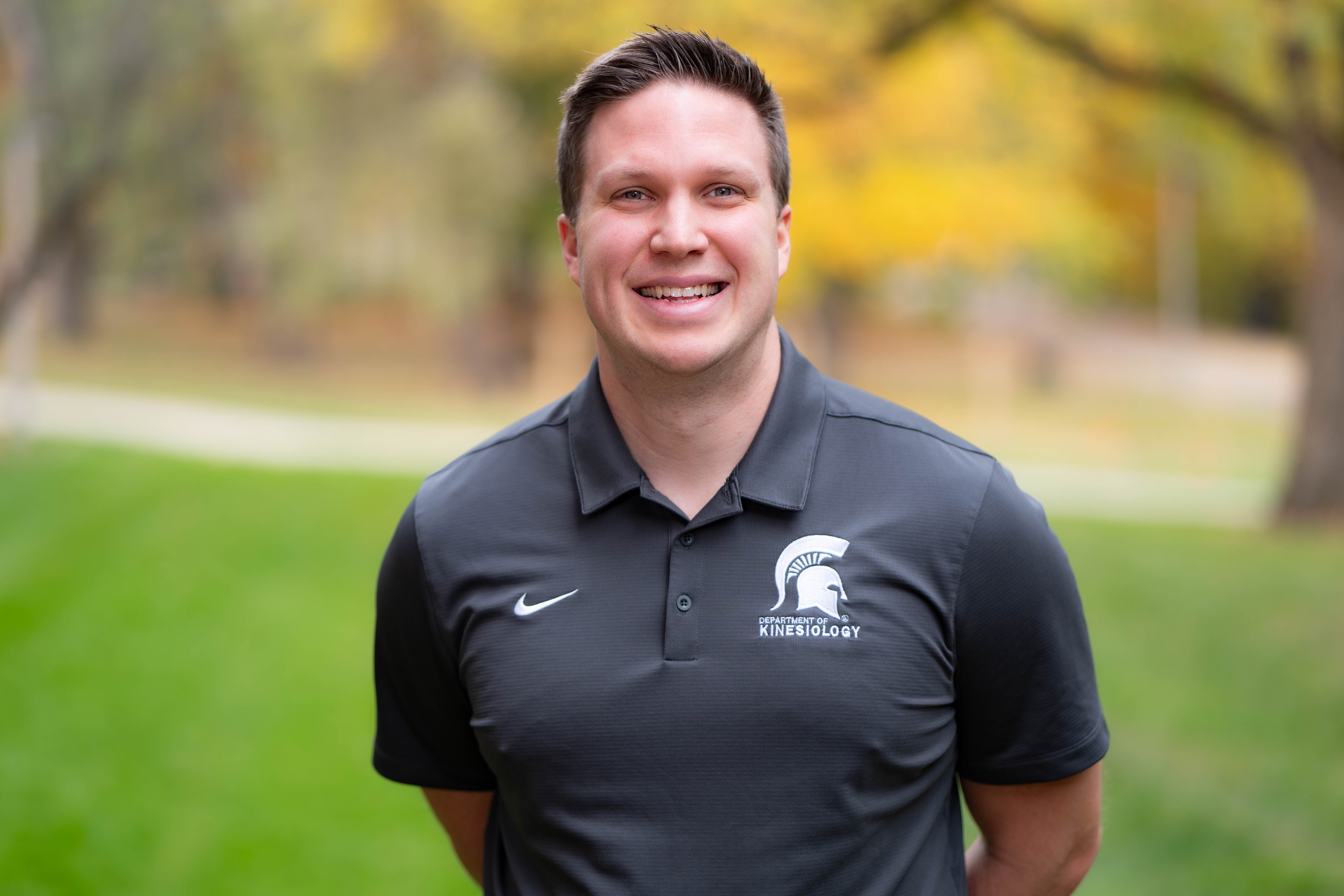 Nicholas Mortensen wearing a Kinesiology polo. There is fall foliage in the background.