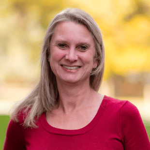 Carey Ommen wearing a red sweater. There is fall foliage in the background. 