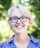 Lynn Paine headshot. Lynn smiles for the camera in front of trees lush with green leaves. Lynn wears a blue floral blouse and dark-rimmed glasses. Lynn's blonde hair is cut close to the head.