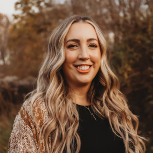 Headshot of Megan Papoi wearing a tan jacket and black shirt, standing in front of fall foliage.