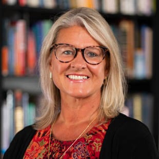 Lisa Plascencia wearing a red top, black jacket and glasses standing in front of a bookshelf 