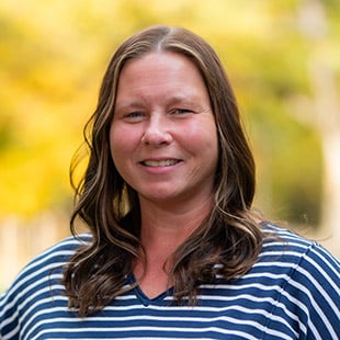 Headshot of Barb Rittenburg wearing a navy striped shirt. She is standing in front of fall foliage