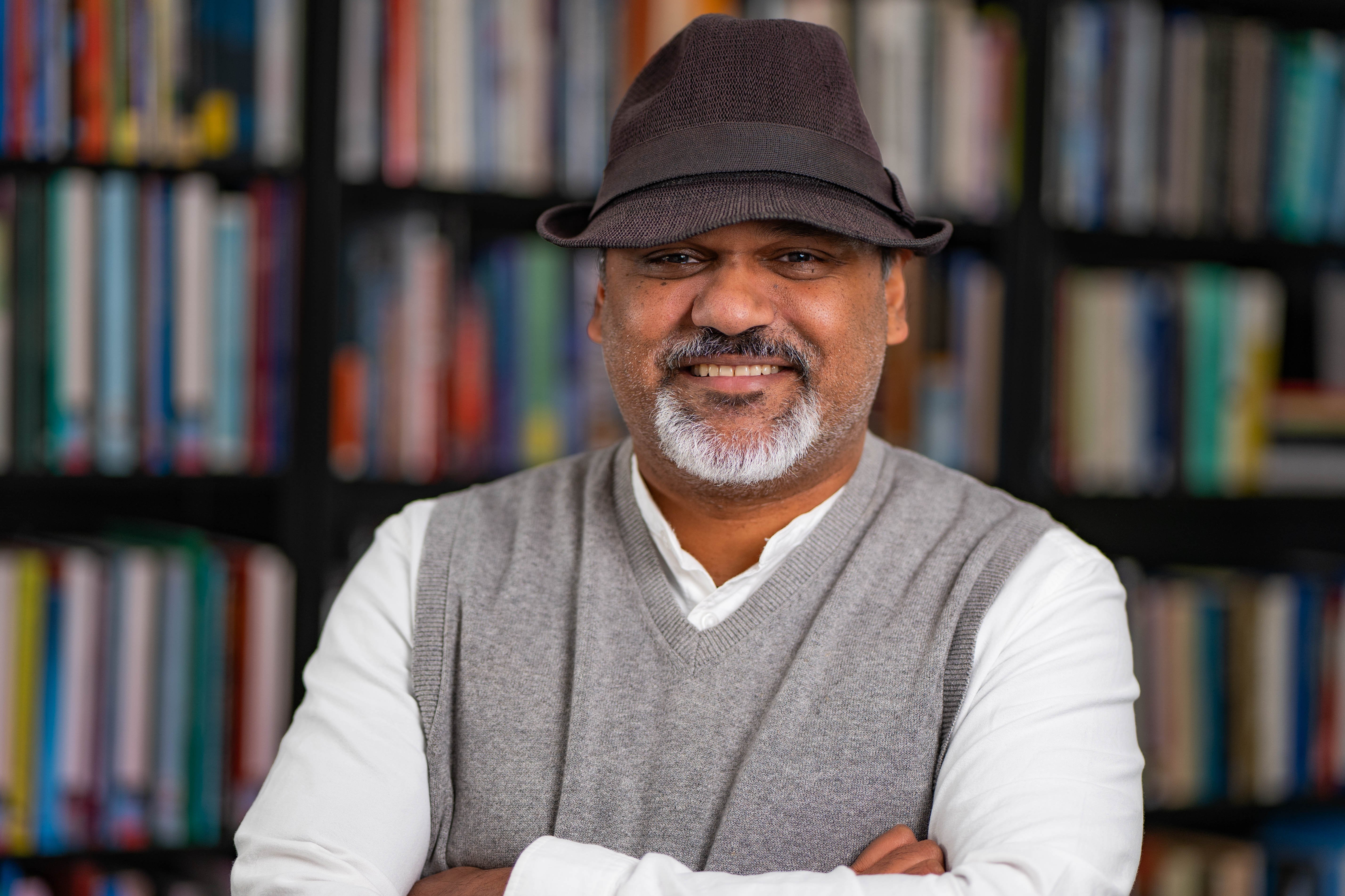 Riyad Shahjahan wearing a white shirt, gray vest and a dark brown hat. There is a bookshelf full of books in the background. 