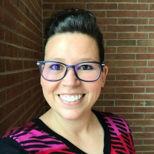Candace Robertson wearing a pink and black shirt with glasses. She is standing in front of a brick wall. 